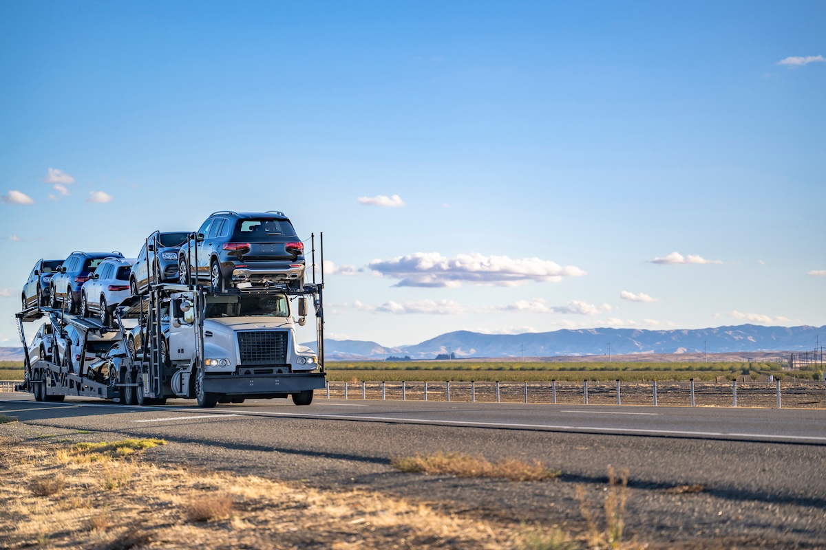 Industrial big rig classic white car hauler semi truck with chrome parts transporting cars on special two level hydraulic modular semi trailer running on the highway road at twilight in California