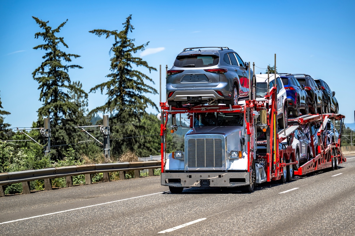 Industrial carrier powered day cab big rig white car hauler semi-truck tractor transporting cars and crossovers on hydraulic two level red semi trailer driving on the wide highway road