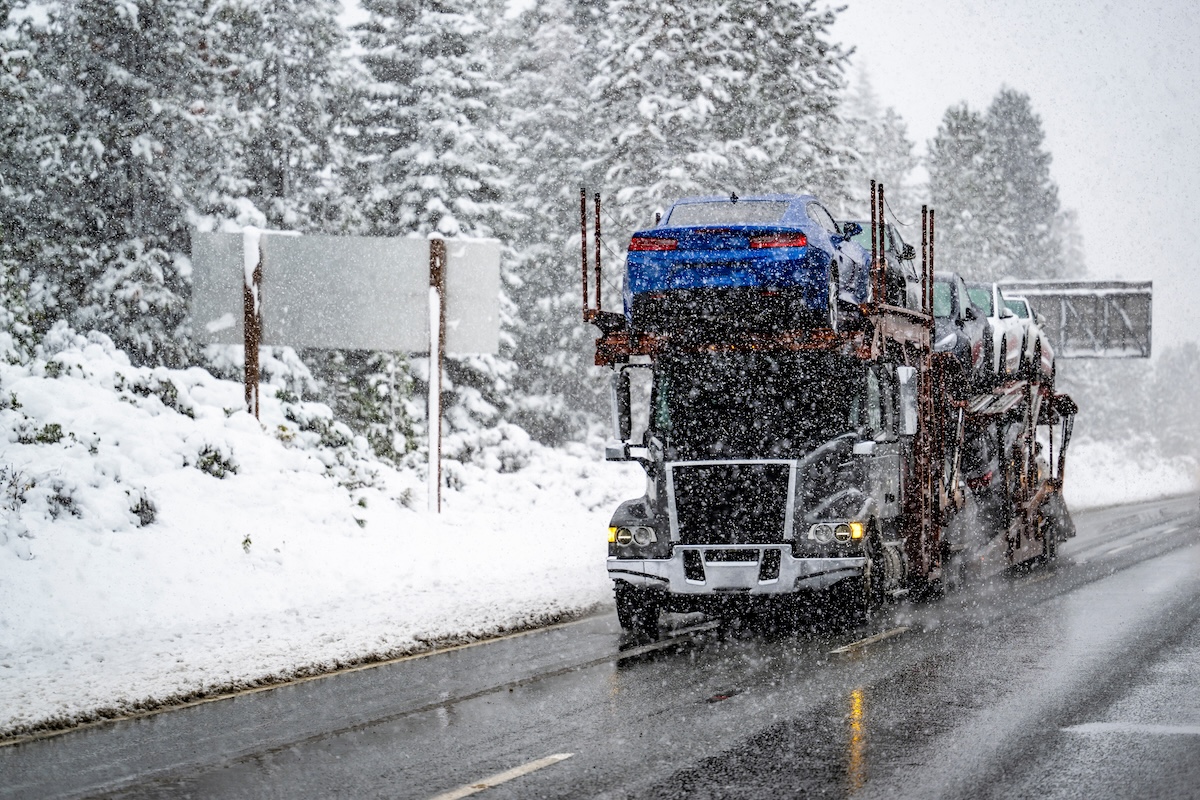 Industrial black big rig car hauler semi truck transporting cars on two level hydraulic semi trailer cautiously driving on a dangerous winter highway during snowfall in Shasta Lake area in California